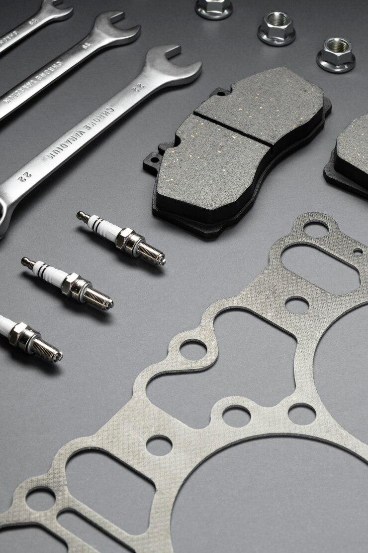 Flat lay of automotive parts including wrenches, brake pads, spark plugs, gaskets, and nuts on a dark gray surface.